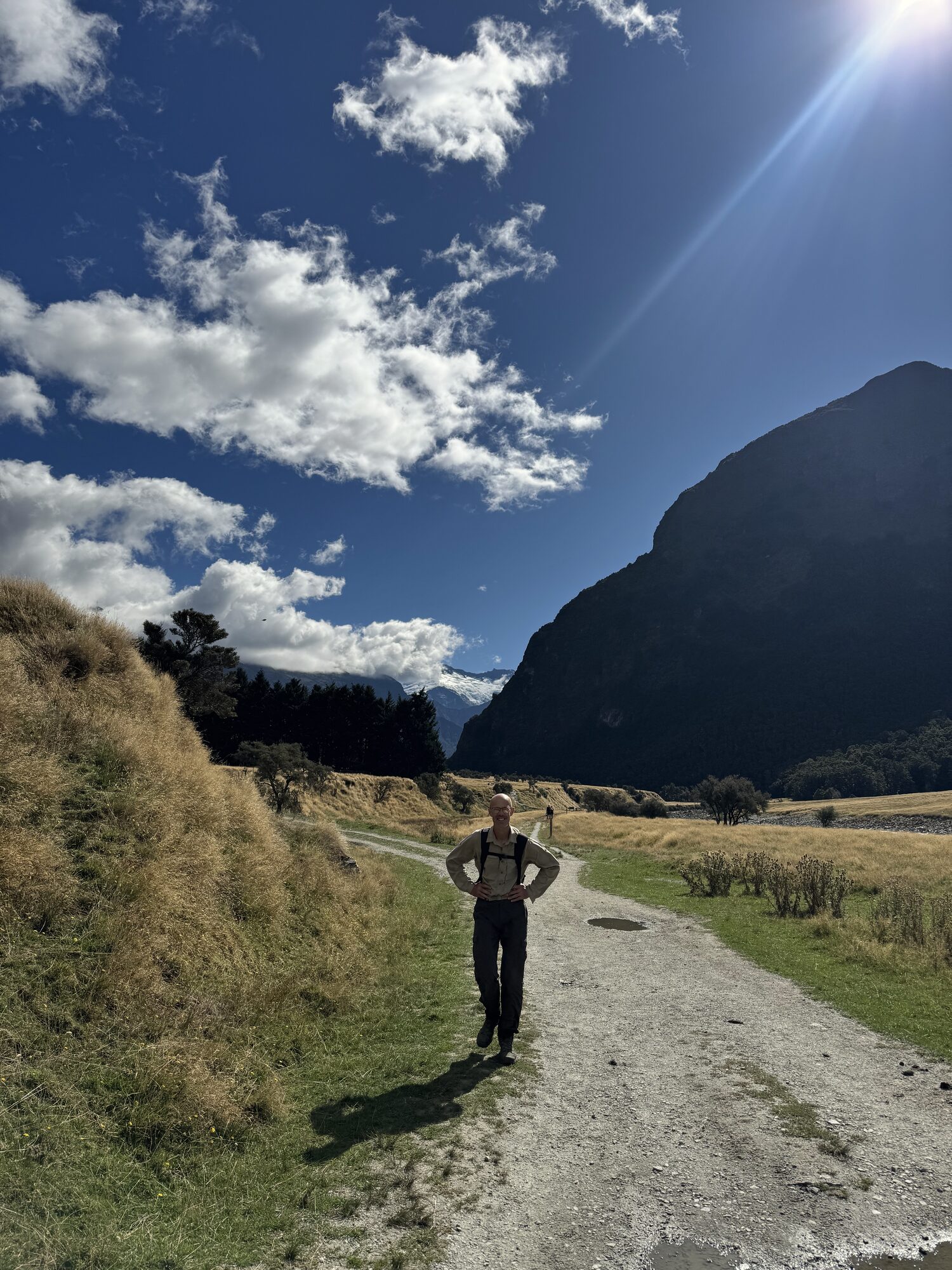 Vic standing on a gravel track in a sunny mountain valley, hands on hips; two walkers are further along the track behind him.