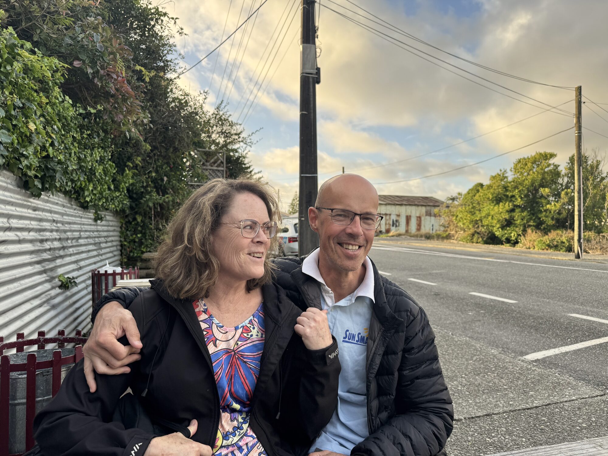 Vic smiling with his arm around a woman in a floral top, both in black puffer jackets, beside a quiet road at dusk.