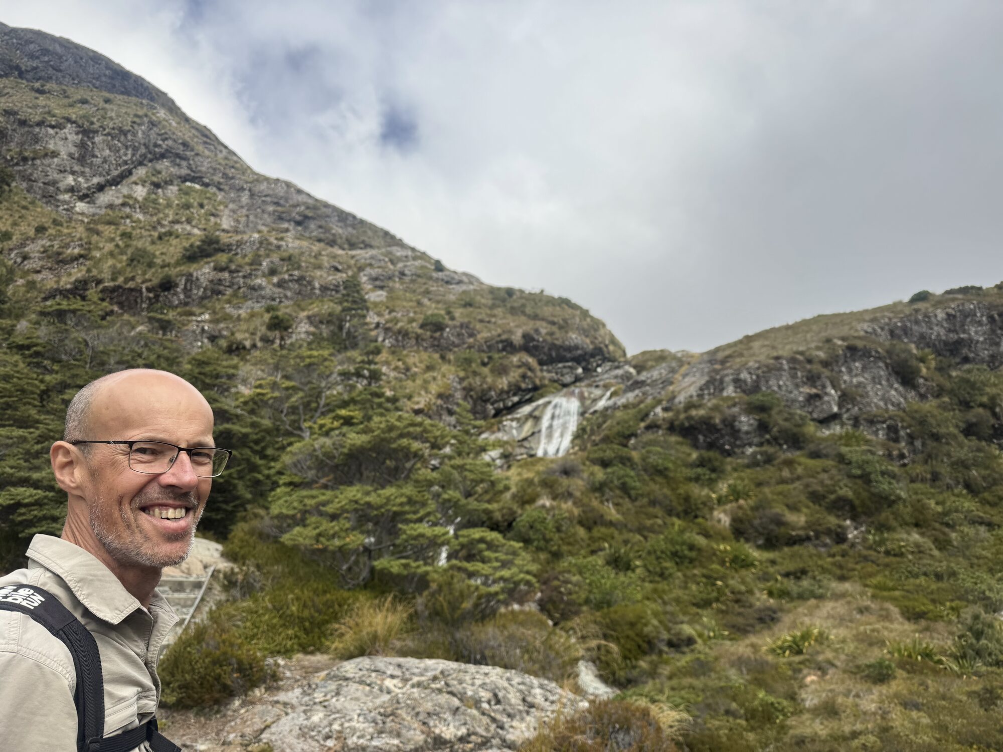 Vic smiling in a selfie on an alpine track, a waterfall and tussock slopes behind him.